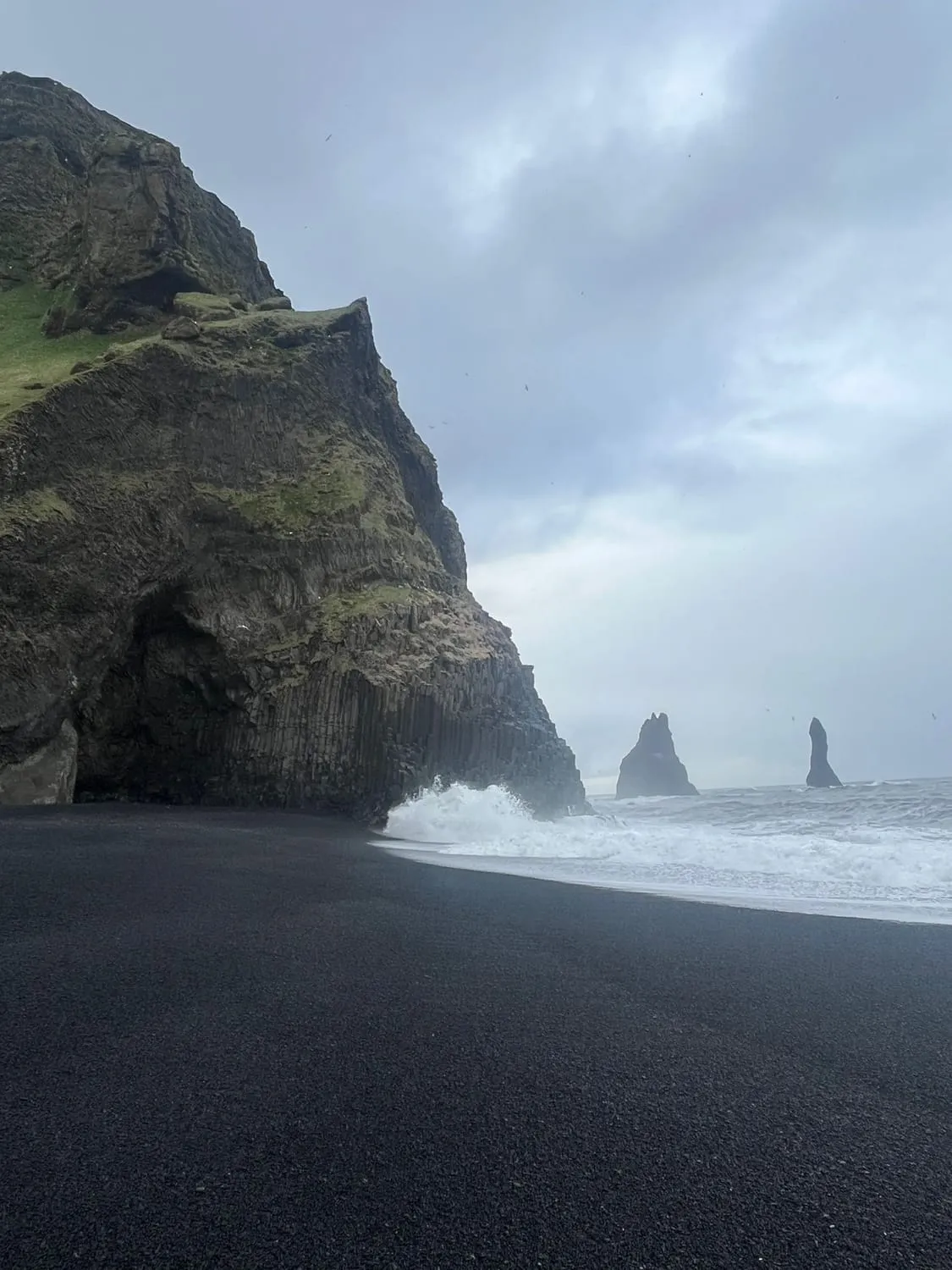 A black beach below a dark cliff, sea stacks in the mist beyond.