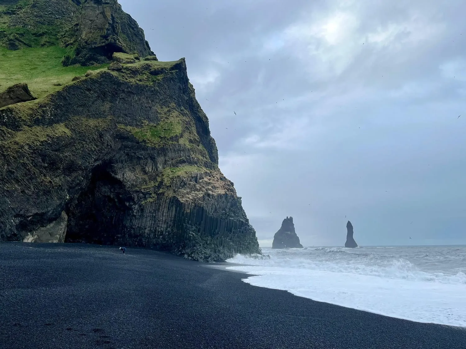 A black sand beach curving towards a headland, with sea stacks standing offshore.