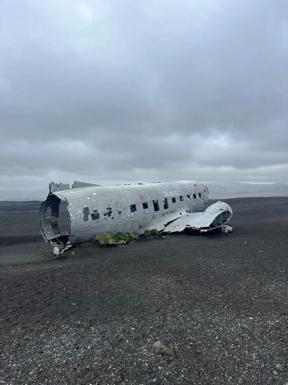 The stripped fuselage of a small aircraft on black sand under a grey sky.