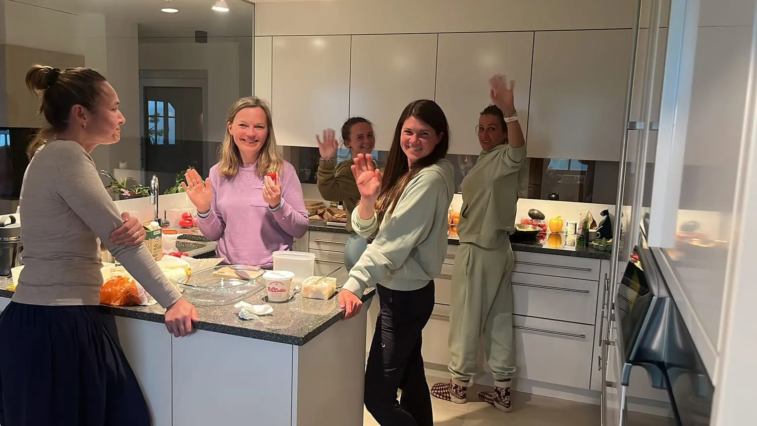 Four people around a kitchen island, food laid out, everyone turning to the camera.