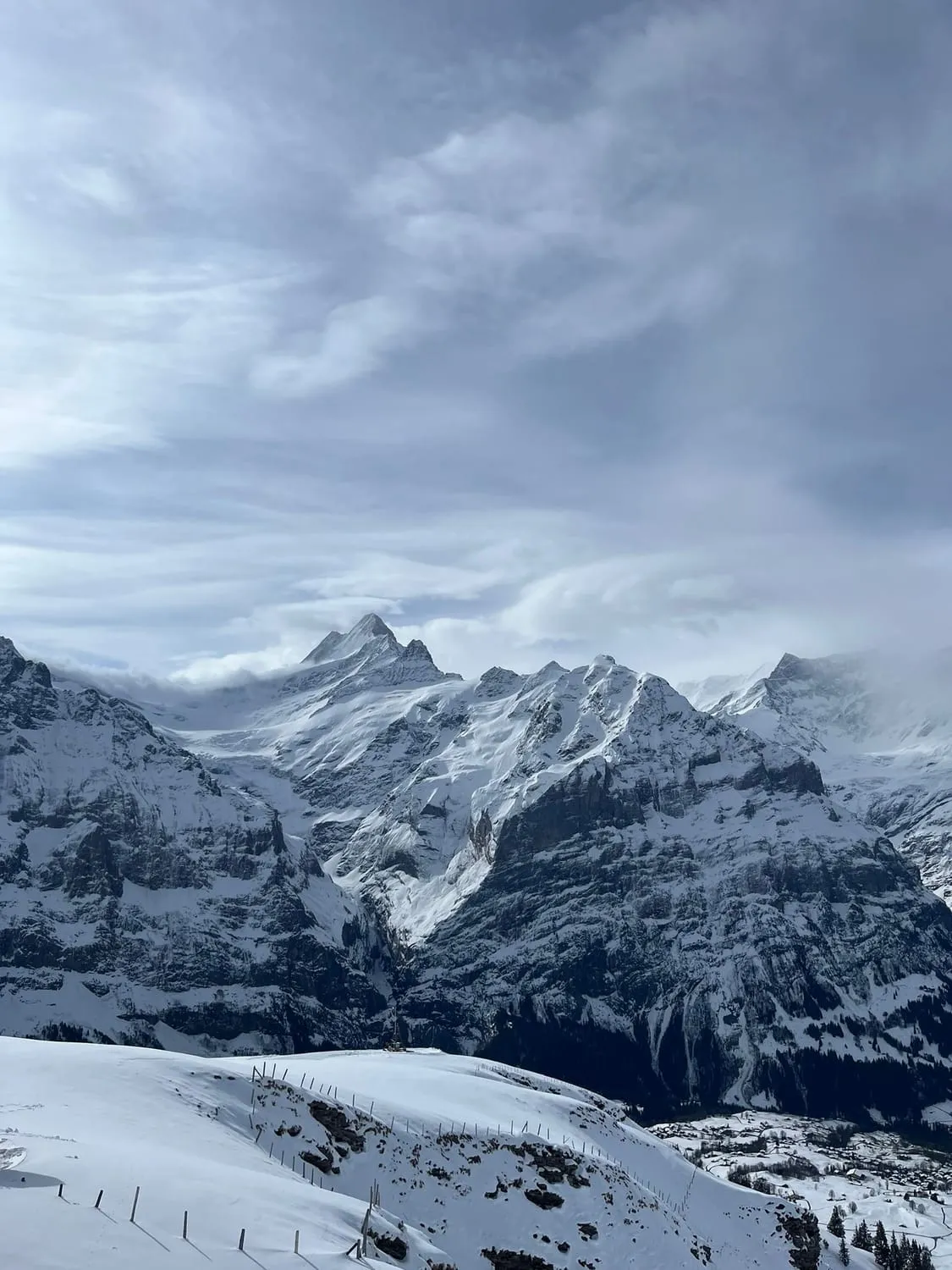 Snow covered peaks under heavy grey cloud, a fence line in the foreground.