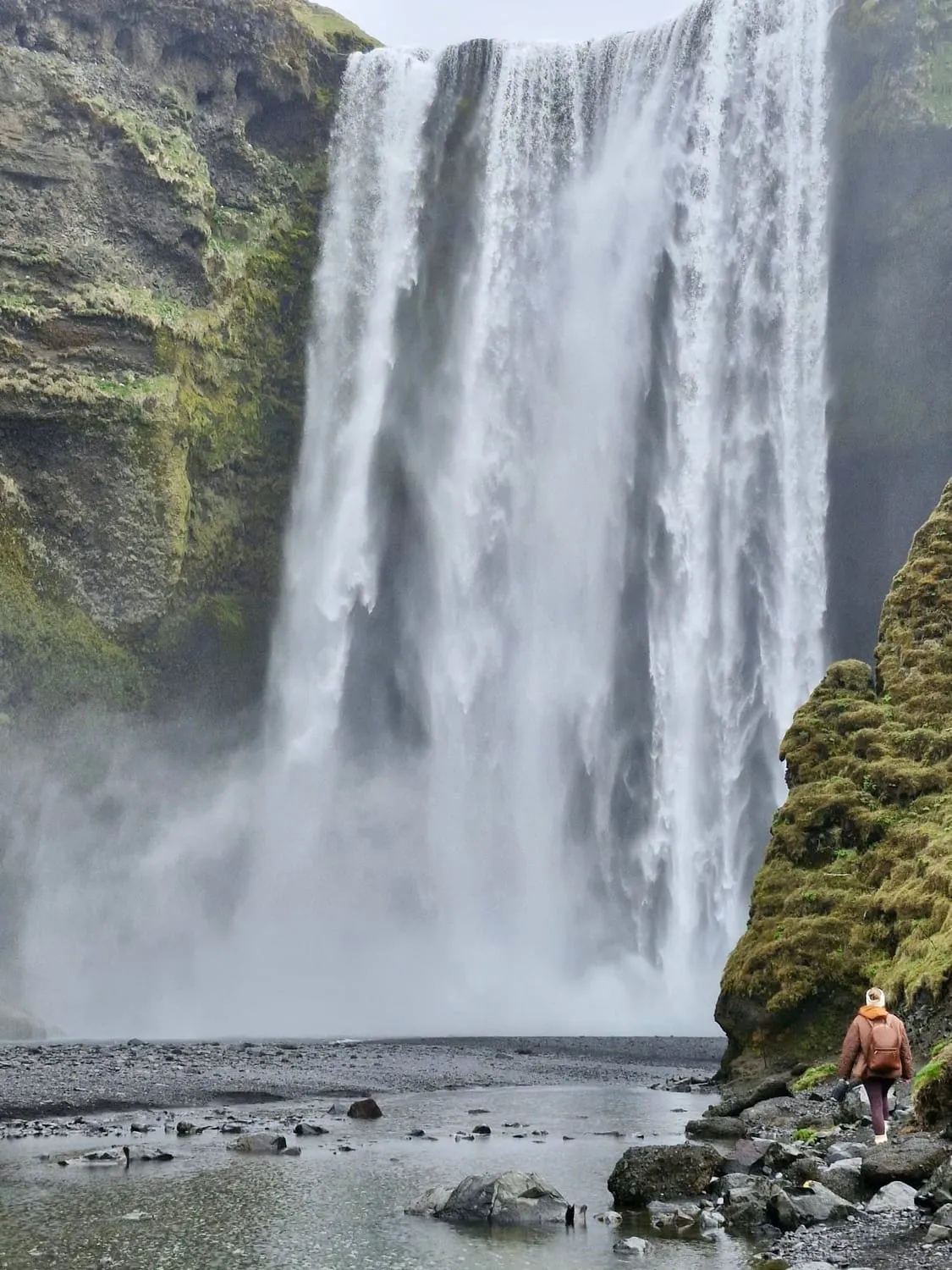 A wide waterfall falling from a green cliff, a small figure standing on the rocks below.